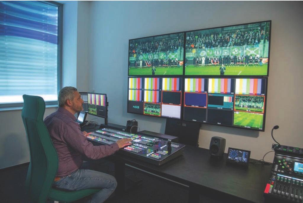 A man sits with a mixing desk in front of him and a large screen attached to the wall, showing a sports game