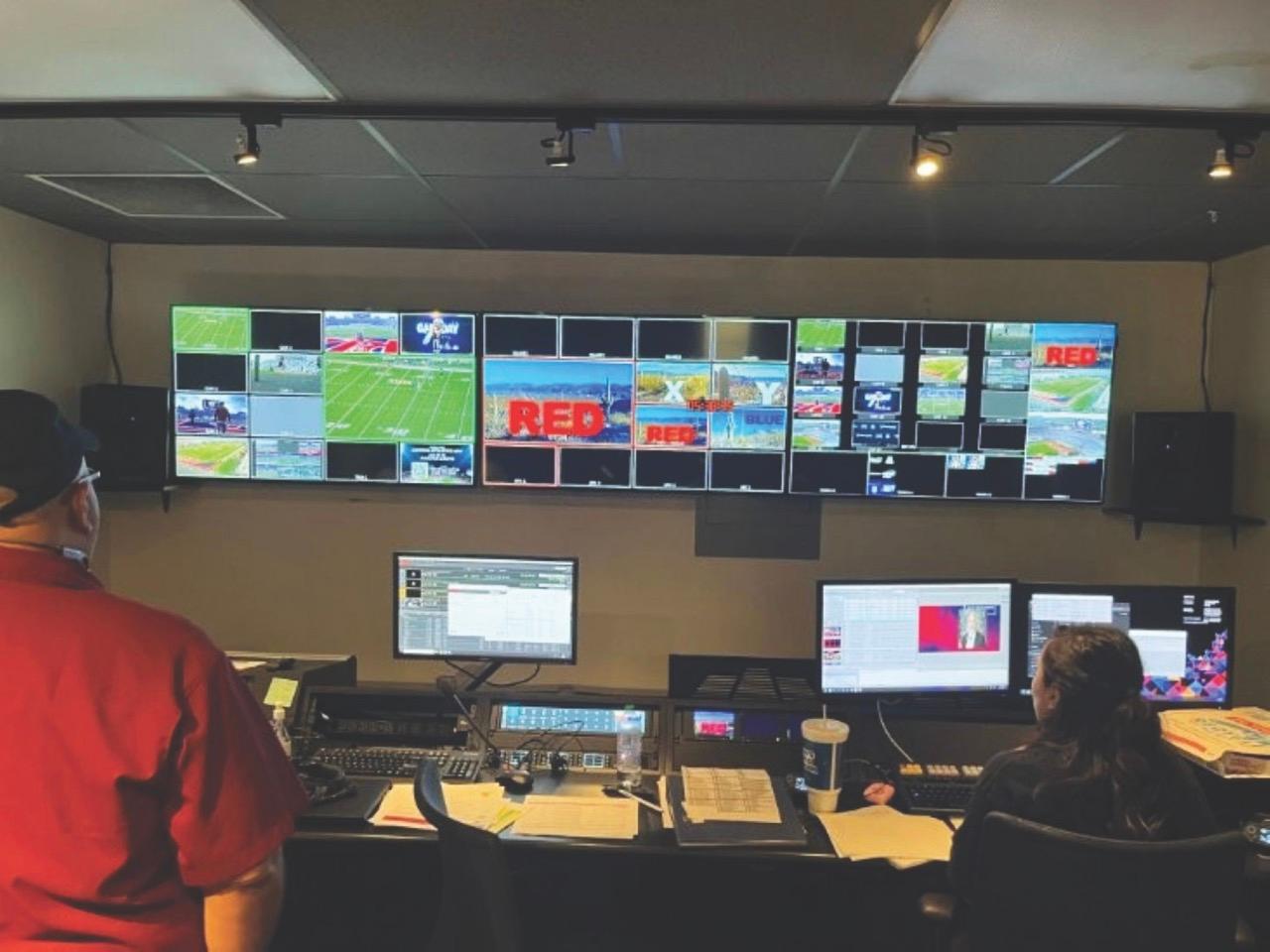 A mxinging room with people working at monitors and looking at screens on a wall, which show a sports game