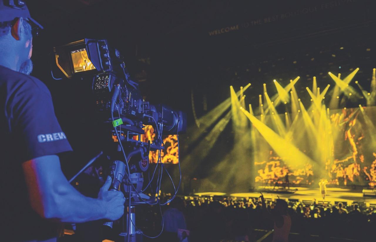 Over the shoulder of a man operating a camera at the back of a concert with a stage lit in yellow light