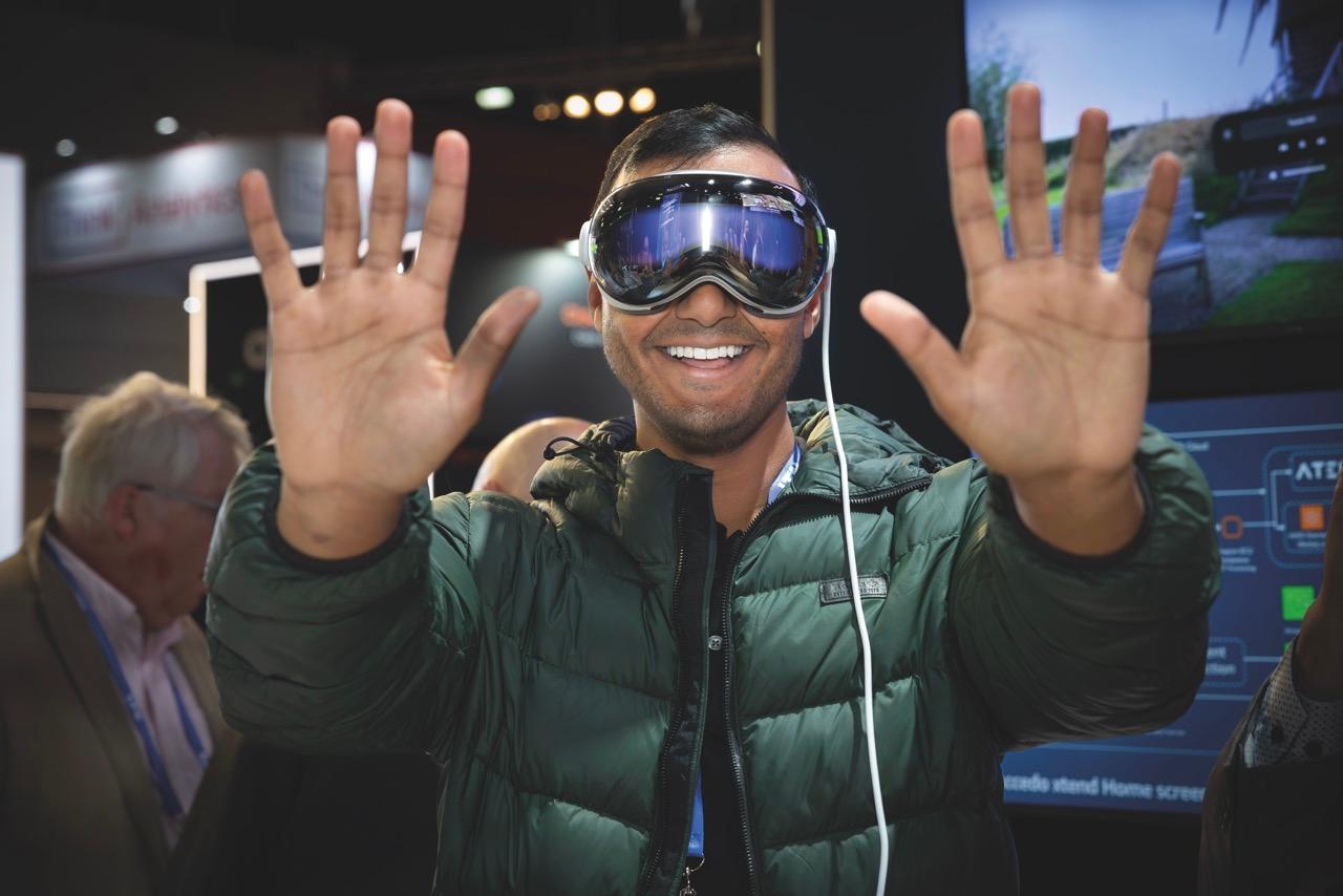 A man wearing a VR headset holds up his hand while another man directs him in the foreground at IBC event