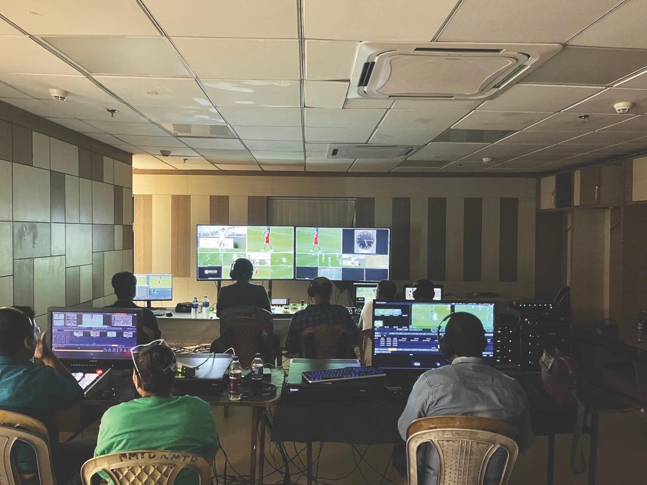 A classroom with people sitting around computers and looking at a football game on a screen at the front