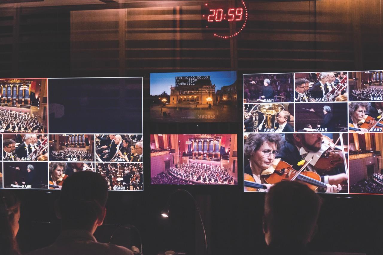 A dark mixing room where people are sitting and watching screens showing an orchestra performance