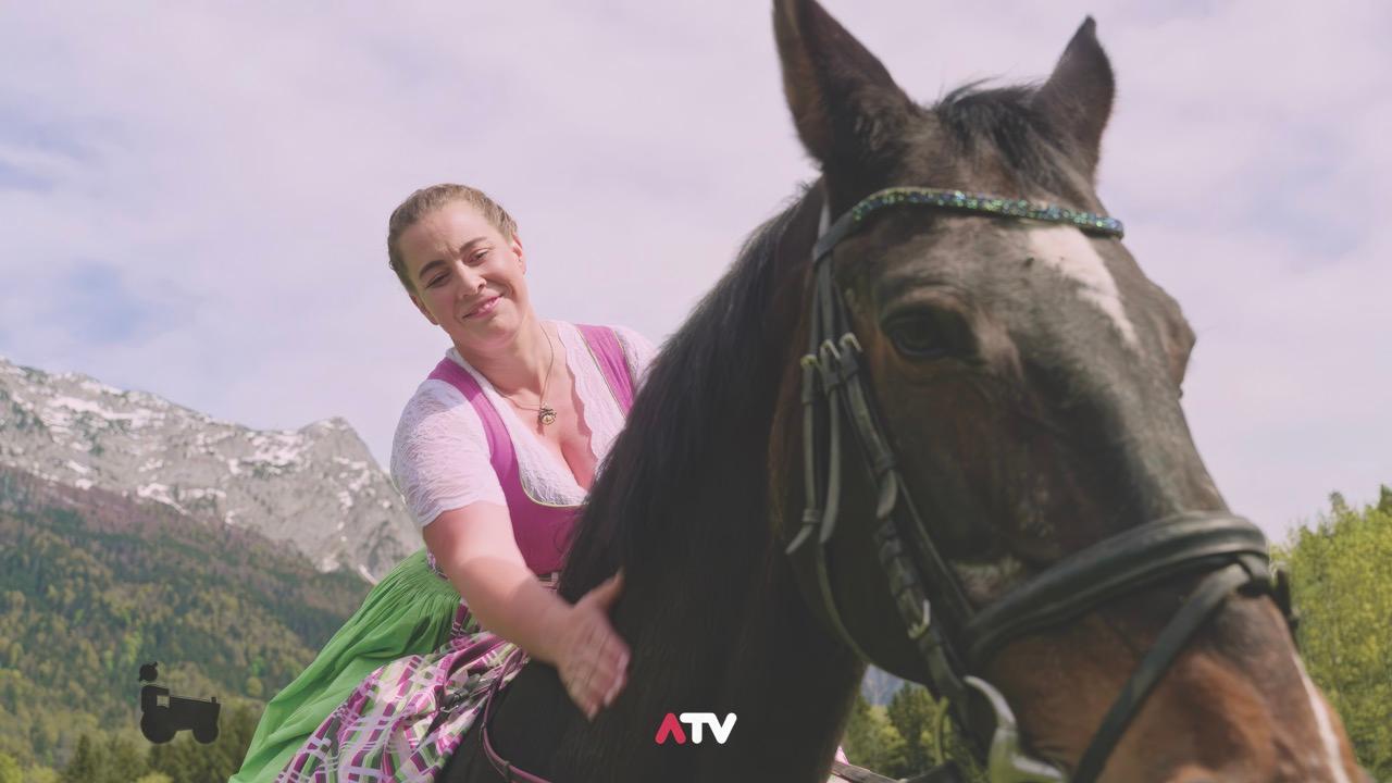 A woman in pink sits on the back of a horse and pats it in a mountainous region
