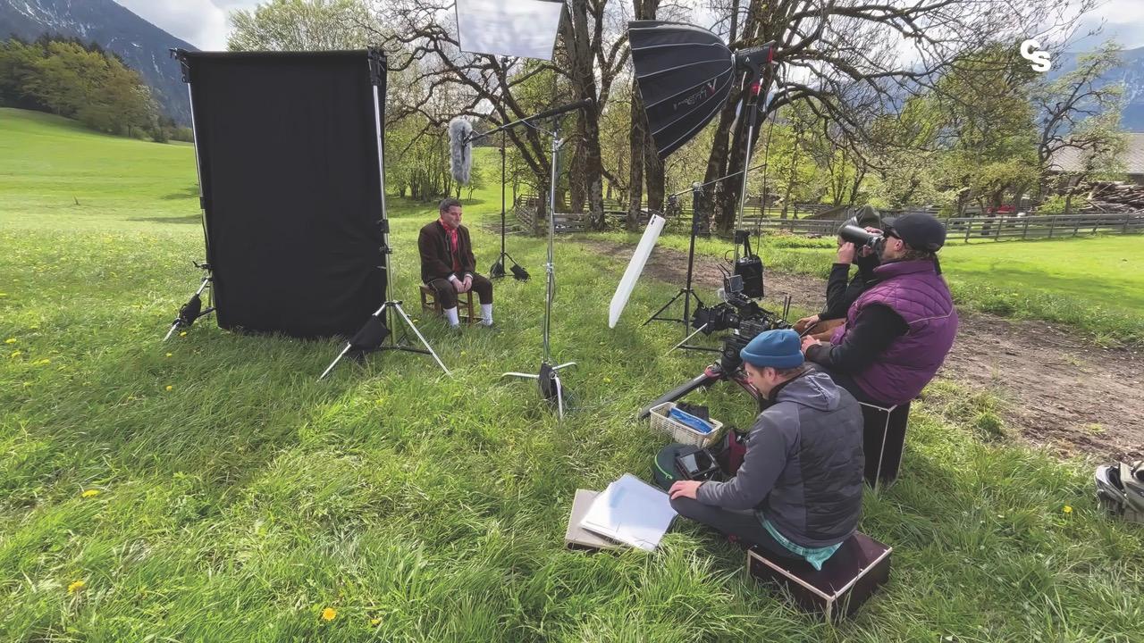 A filming crew sits in a grass field, filming a man sitting next to a tree