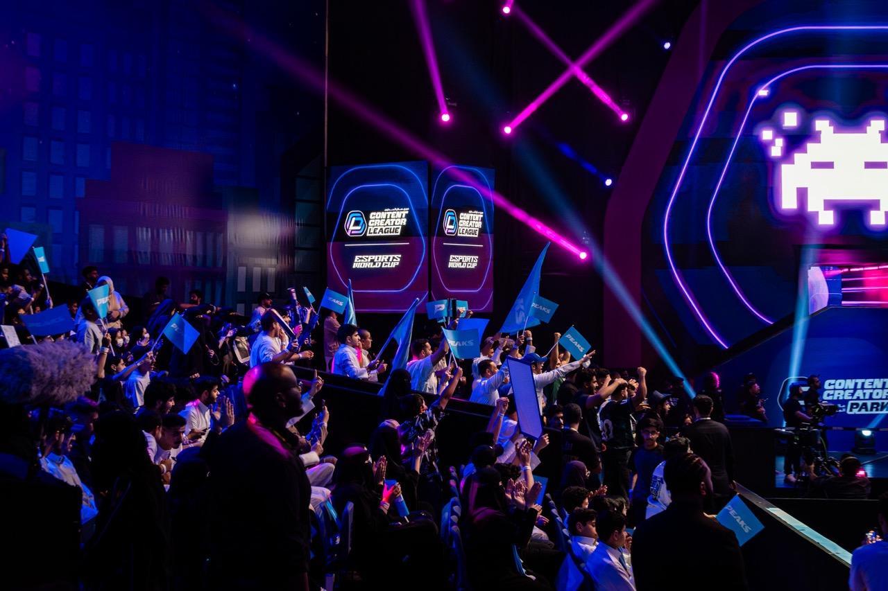 A large audience holding up banners at the Esports World Cup with screens showing vintage games