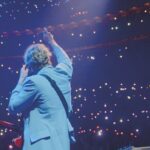 Behind a man in a blue suit holding a guitar performing in an arena