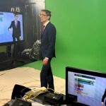 A person in a suit standing in front of a green wall presenting the weather. He stands on a brown cloth with a TV next to him