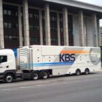 A large white KBS truck on the street in front of a big building