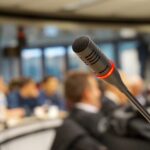 A close-up of a microphone in a filled conference room with a blurred background