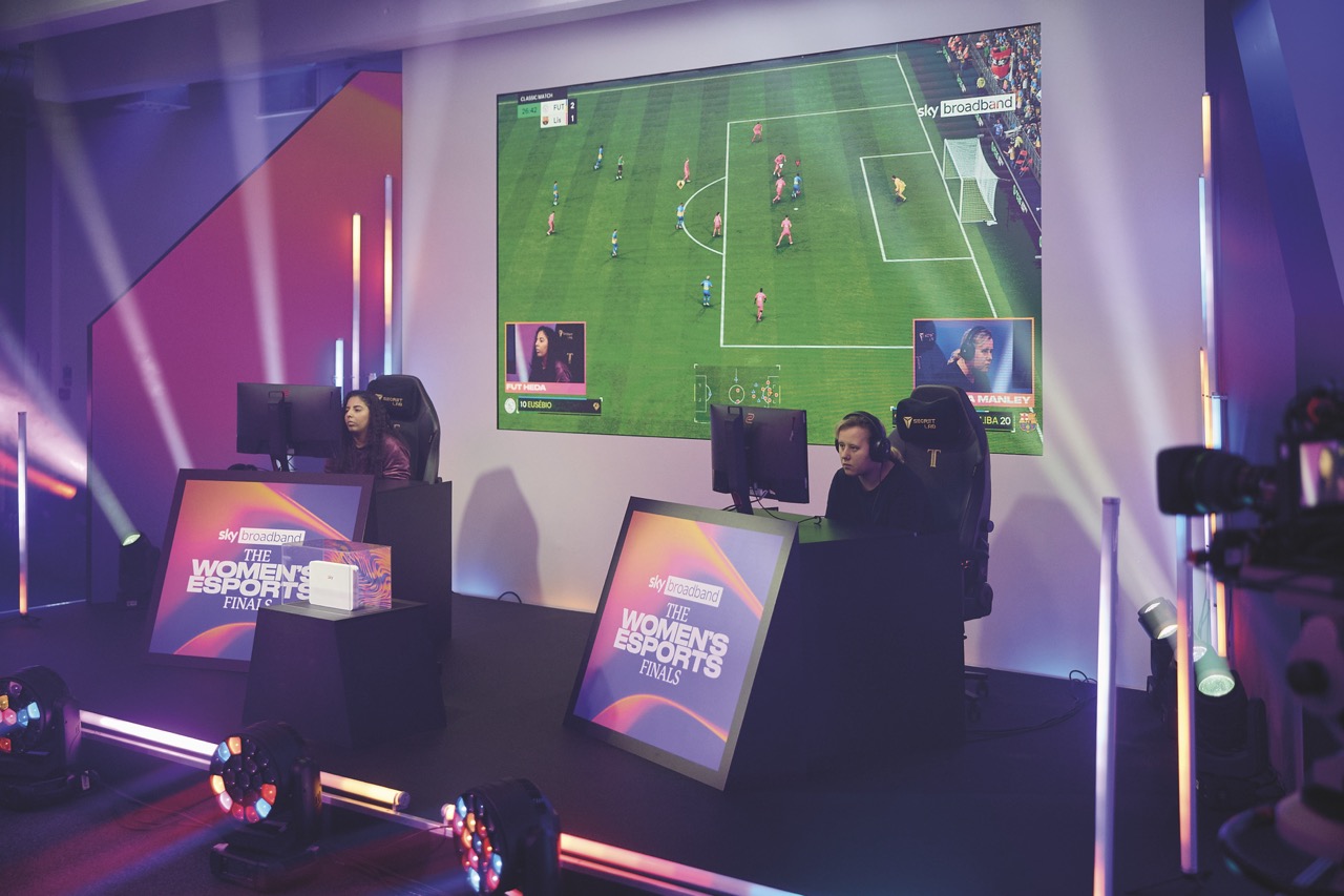 Two women sitting at gaming chairs. There is a large LED screen behind them showing a football game