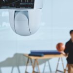 A white camera hanging upside down from the ceiling, filming a person throwing a basketball in a blue room